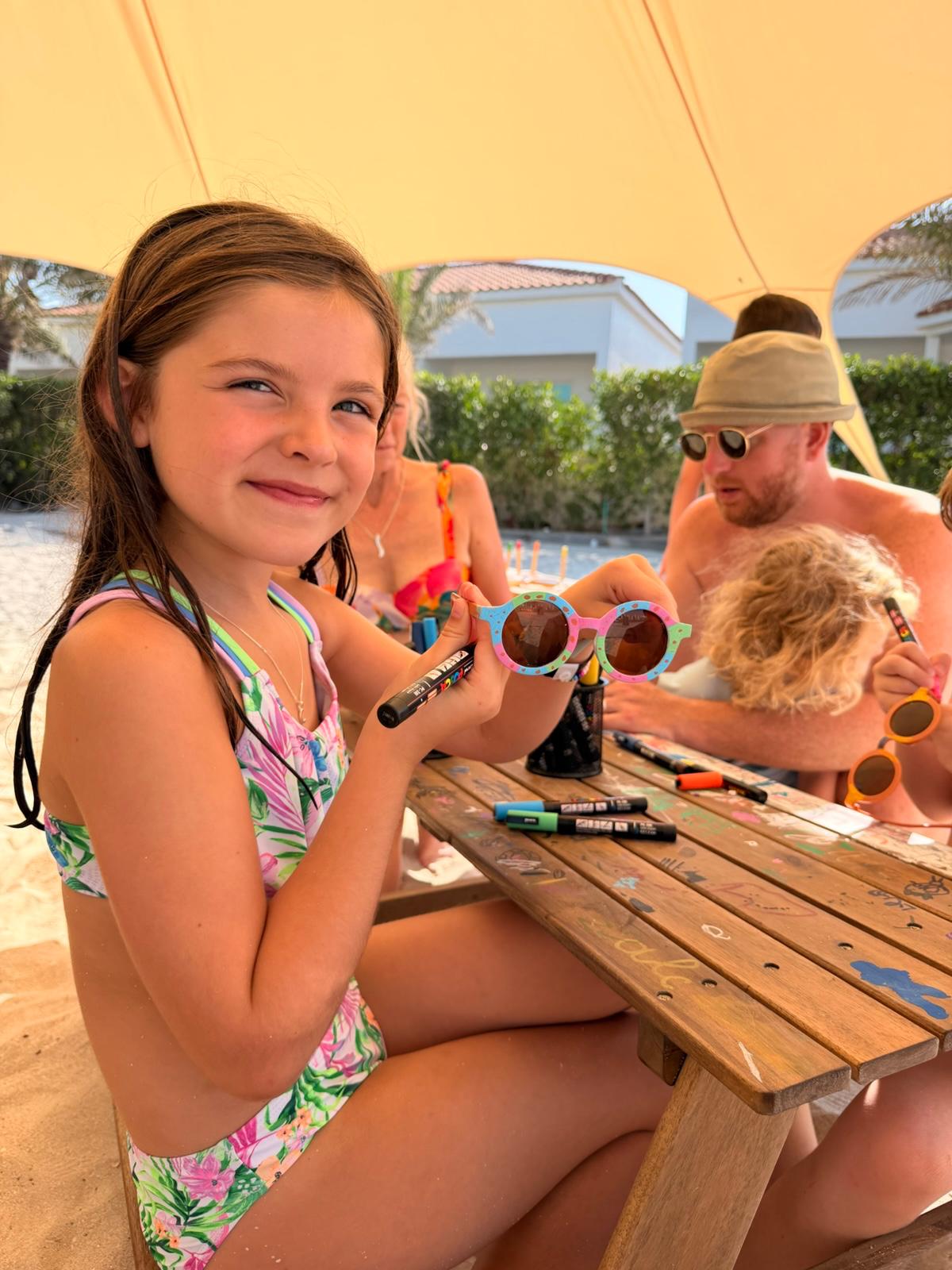 Children decorating sunglasses together at a shaded beach table